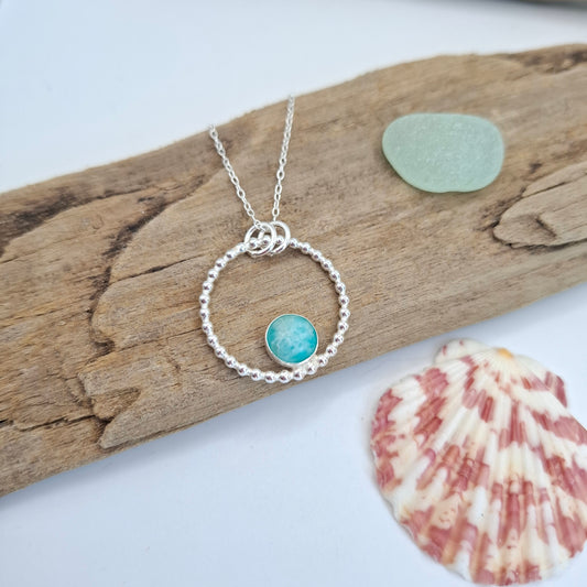 A silver necklace with a textured circular ring and a small round turquoise-blue stone charm, displayed on a piece of driftwood with a sea glass pebble and a seashell nearby.