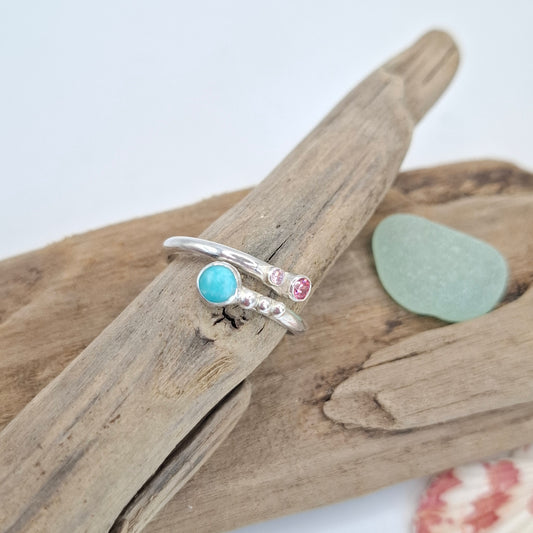 Two silver rings displayed on a piece of driftwood. One ring features a round light blue stone, and the other has a small round pink stone and tiny clear stones.