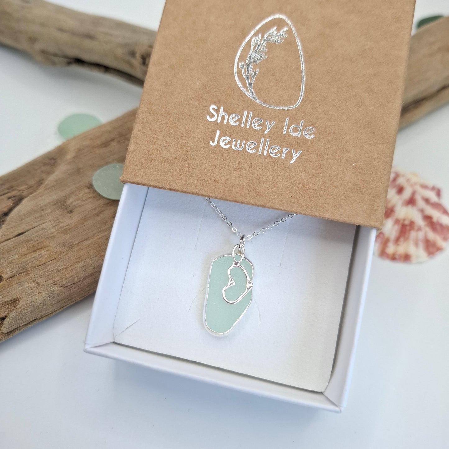 A silver necklace with a light green sea glass pendant is displayed in an open white gift box next to its brown kraft paper lid, which is printed with the brand name 'Shelley Ide Jewellery'.