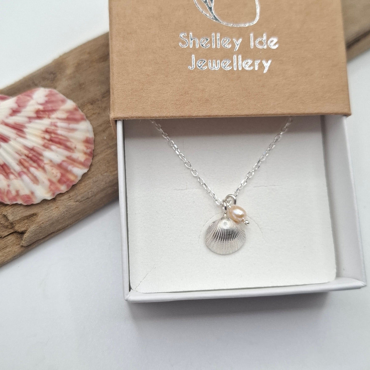 A silver shell pendant necklace with a small pearl charm, presented in an open white jewelry box with a brown kraft lid printed with 'Shelley Ide Jewellery'. A pink and white seashell and a piece of driftwood are styled on the left side.