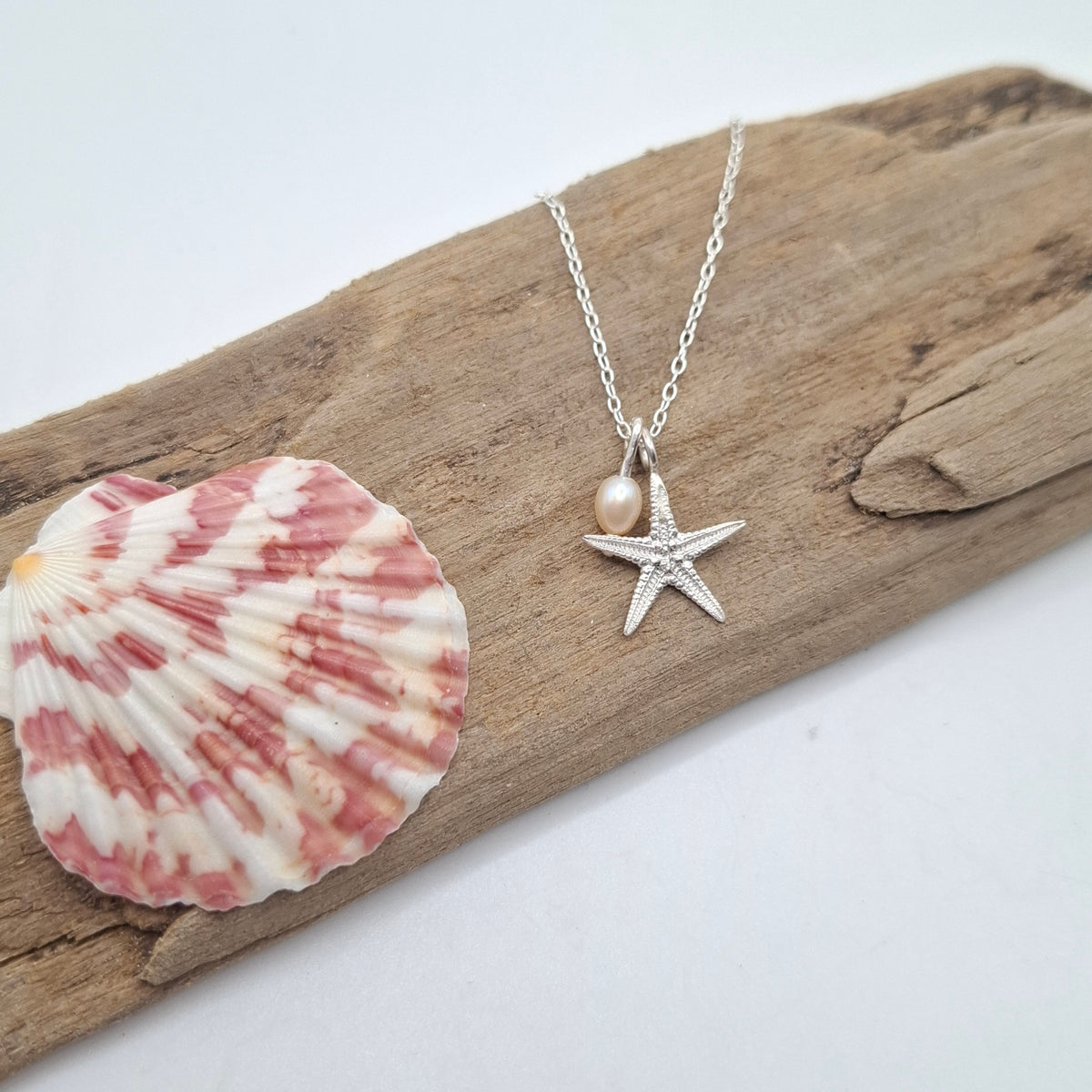 A silver starfish pendant necklace with a small pearl bead, displayed on a piece of driftwood next to a pink and white seashell against a white background.