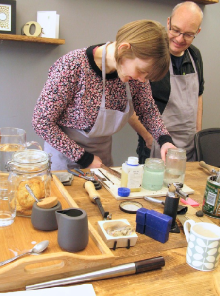 A woman in a floral long-sleeve top and white apron is leaning over a wooden worktable, mixing or pouring something into a glass jar. A man in a dark shirt and gray apron stands behind her.