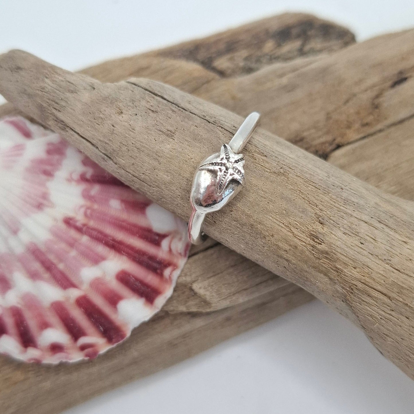 Silver ring with a starfish design on a pink shell and driftwood against a white background