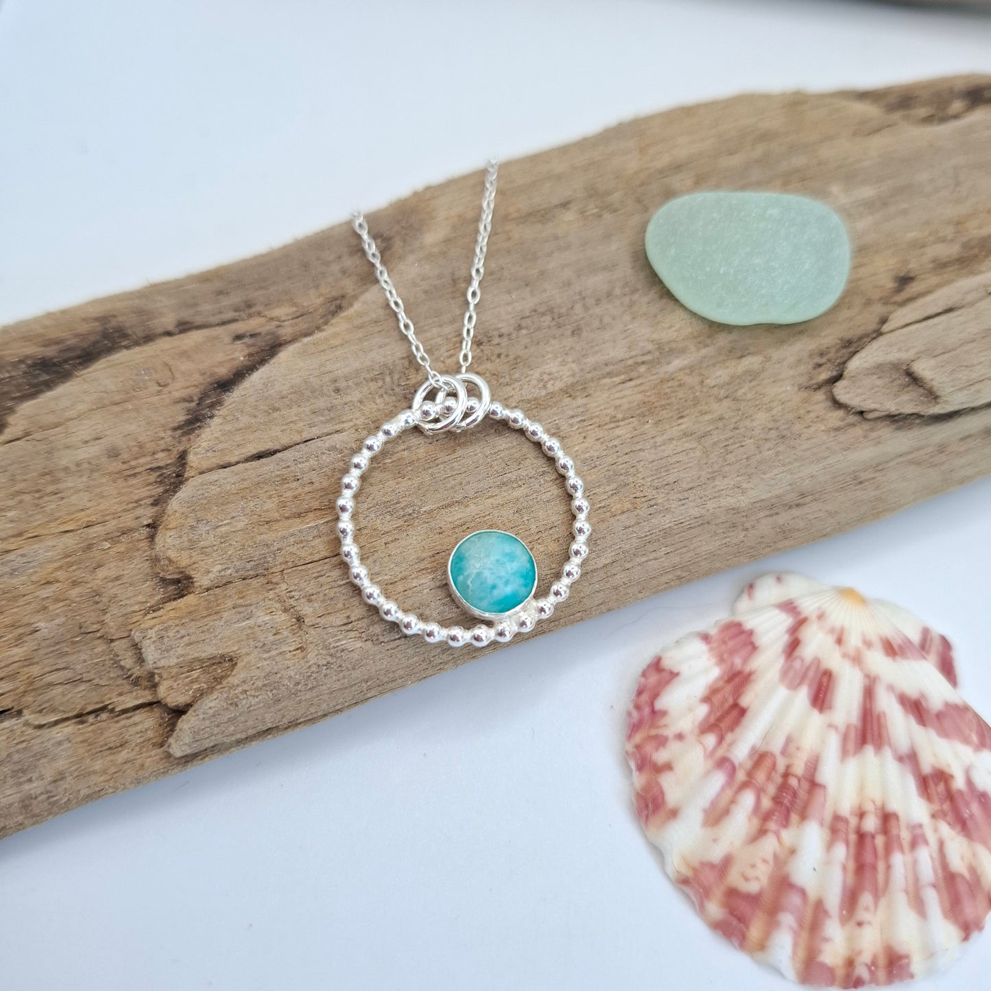 A silver necklace with a textured circular ring and a small round turquoise-blue stone charm, displayed on a piece of driftwood with a sea glass pebble and a seashell nearby.