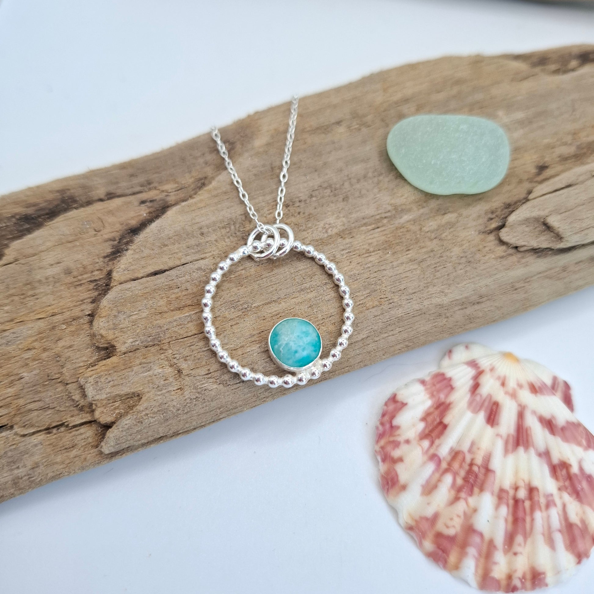 A silver necklace with a textured circular ring and a small round turquoise-blue stone charm, displayed on a piece of driftwood with a sea glass pebble and a seashell nearby.