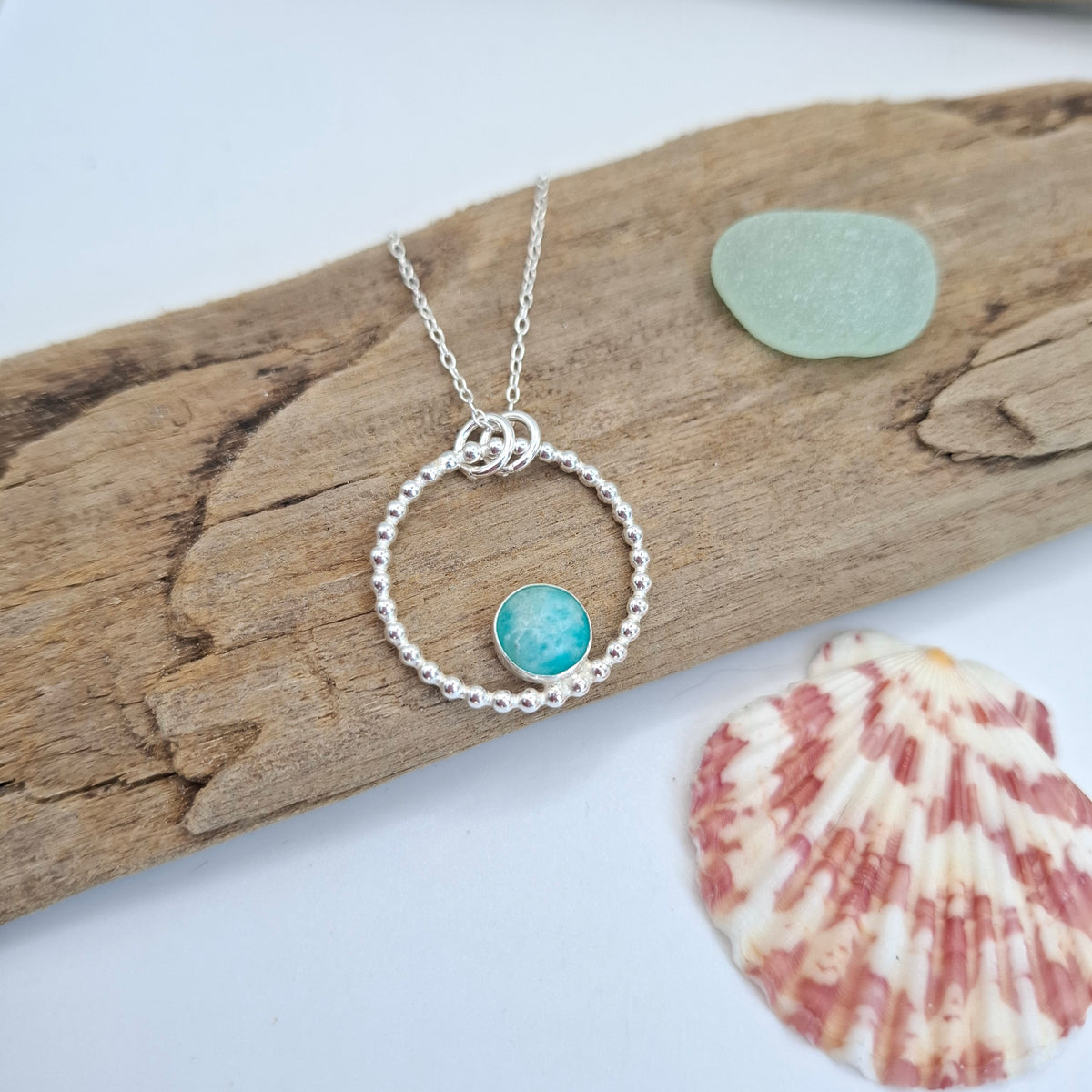 A silver necklace with a textured circular ring and a small round turquoise-blue stone charm, displayed on a piece of driftwood with a sea glass pebble and a seashell nearby.