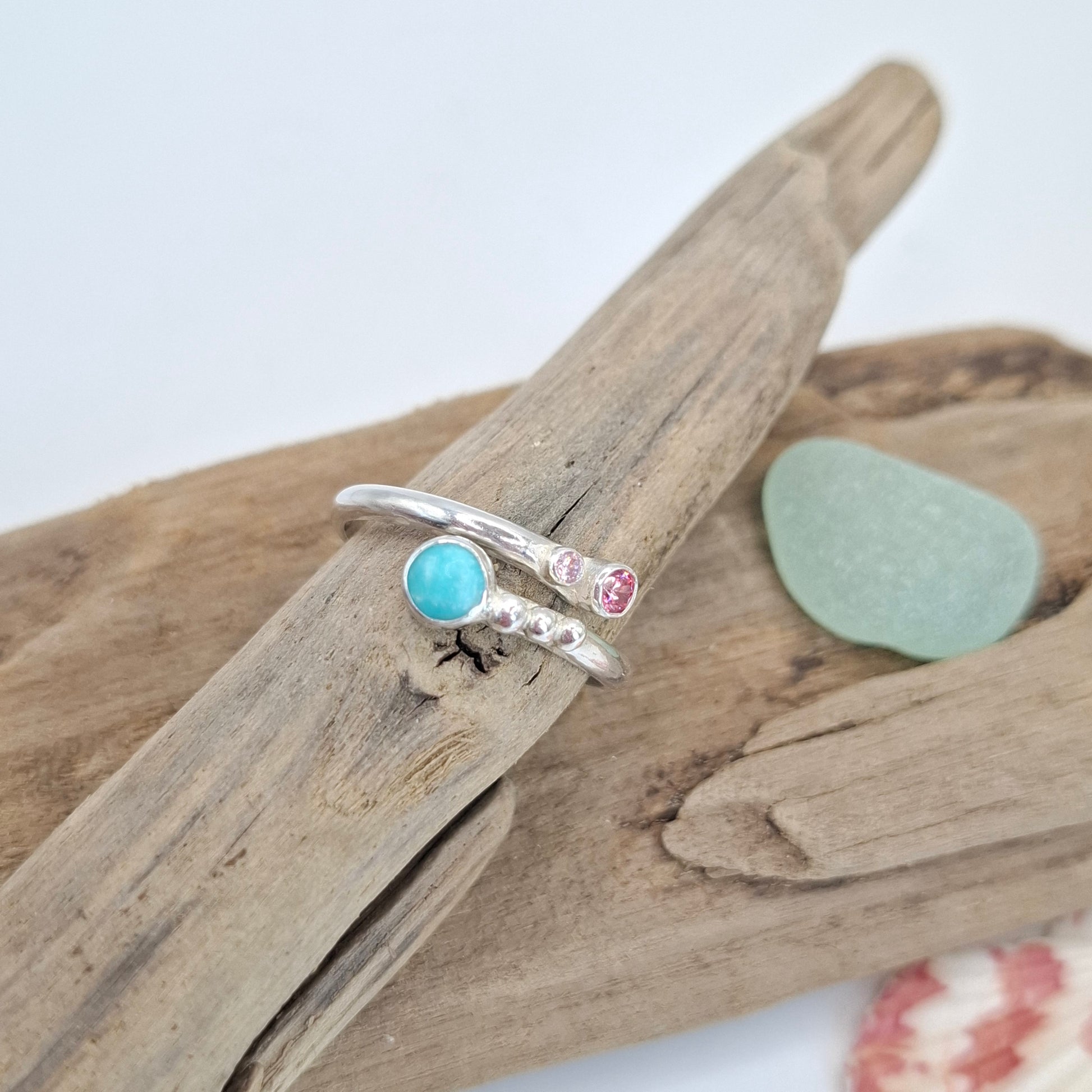Two silver rings displayed on a piece of driftwood. One ring features a round light blue stone, and the other has a small round pink stone and tiny clear stones.