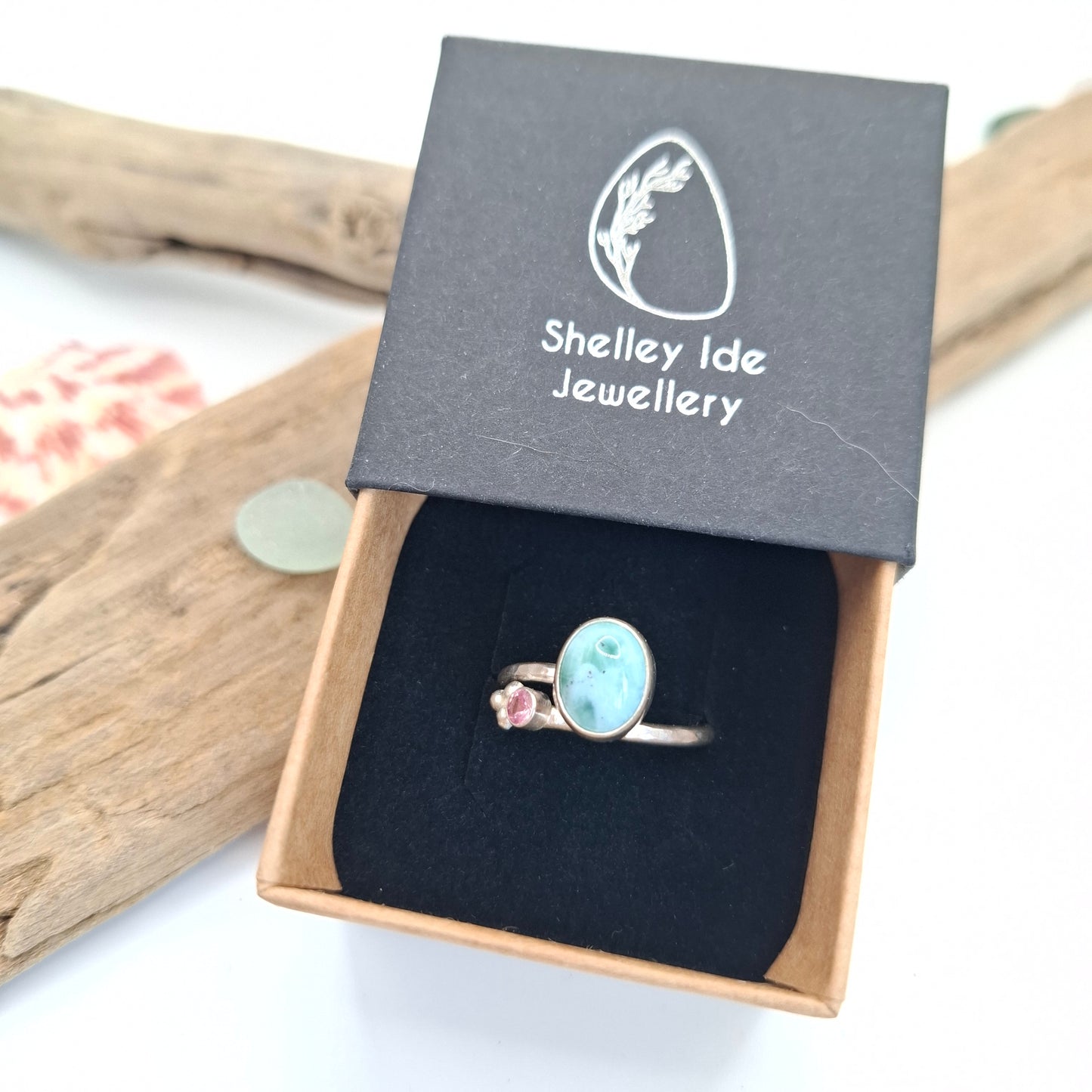 A silver ring with a light blue oval stone and a small pink gemstone, presented in an open brown jewelry box with a black velvet insert. The lid of the box is propped up, showing the brand name 'Shelley Ide Jewellery'.