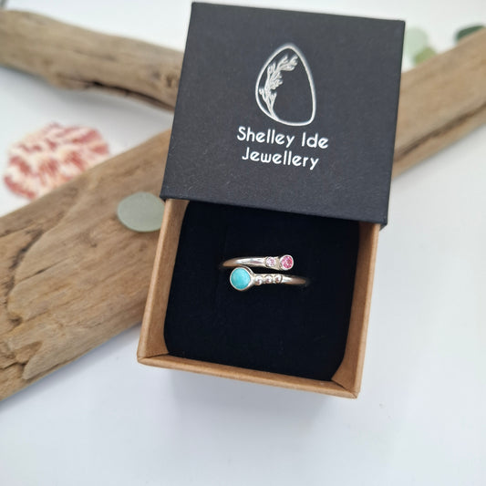 A pair of silver-toned rings displayed in an open black jewelry box with the lid printed with the Shelley Ide Jewellery brand name. One ring features a small pink stone and the other a round turquoise-blue stone, both set in silver bands with small clear stones.