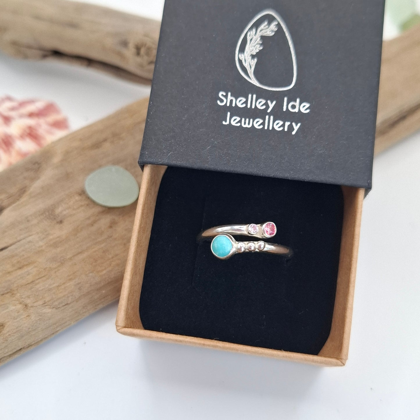 A silver-toned ring with a small pink gemstone and a round turquoise-blue stone, displayed in an open black jewelry box with the brand name 'Shelley Ide Jewellery' visible on the lid.