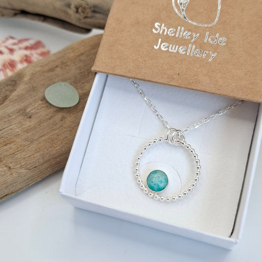 A silver necklace with a circular beaded pendant and a turquoise-colored stone, presented in a white jewelry box next to its kraft paper packaging from Shelley Isle Jewellery.