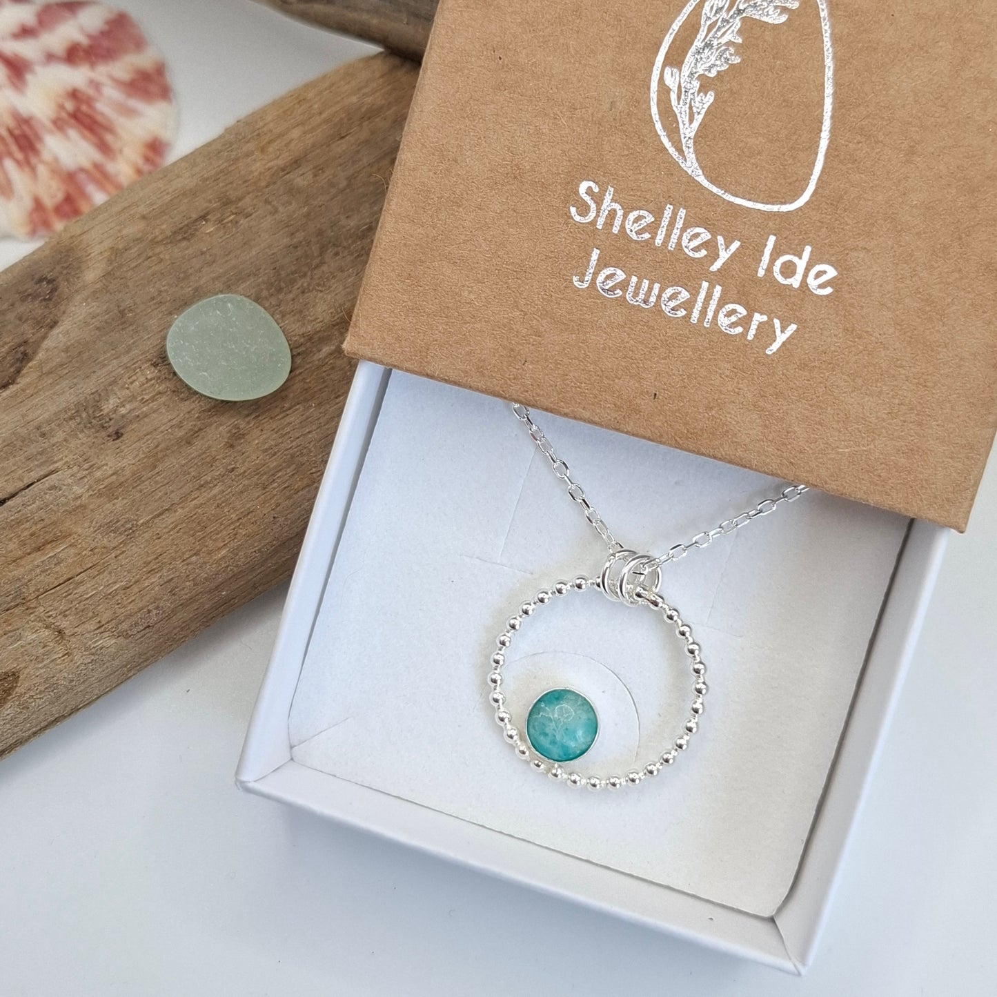A silver beaded necklace with a round turquoise stone pendant, presented in a white jewelry box next to a brown kraft paper box printed with 'Shelley Ide Jewellery'.
