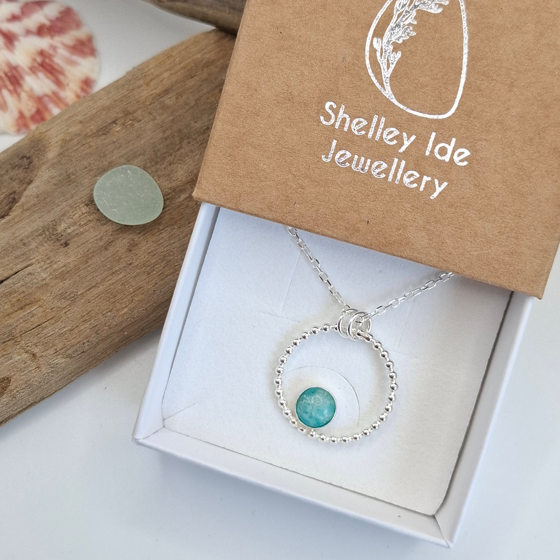 A silver beaded necklace with a round turquoise stone pendant, presented in a white jewelry box next to a brown kraft paper box printed with 'Shelley Ide Jewellery'.