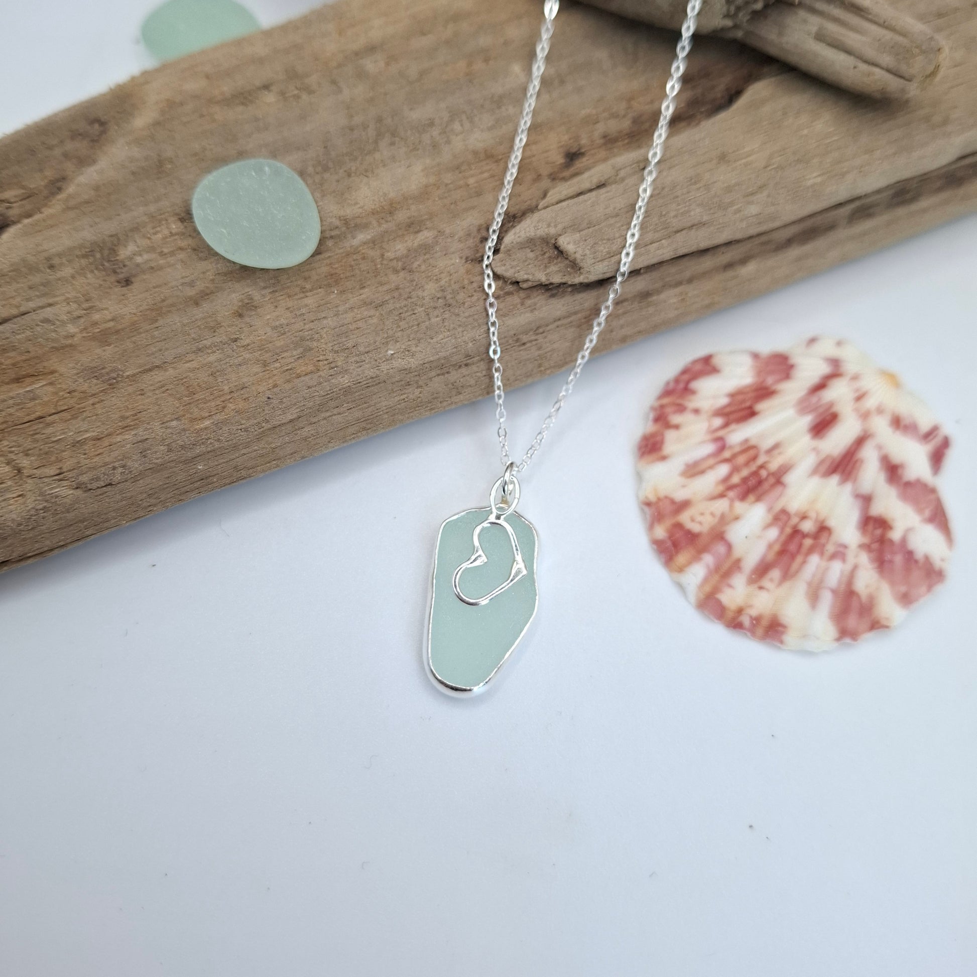 A silver necklace with a sea glass pendant featuring a small heart engraving, styled on a white surface with driftwood, a seashell, and sea glass pieces.