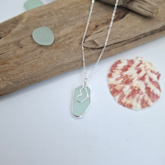A silver necklace with a sea glass pendant featuring a small heart engraving, styled on a white surface with driftwood, a seashell, and sea glass pieces.