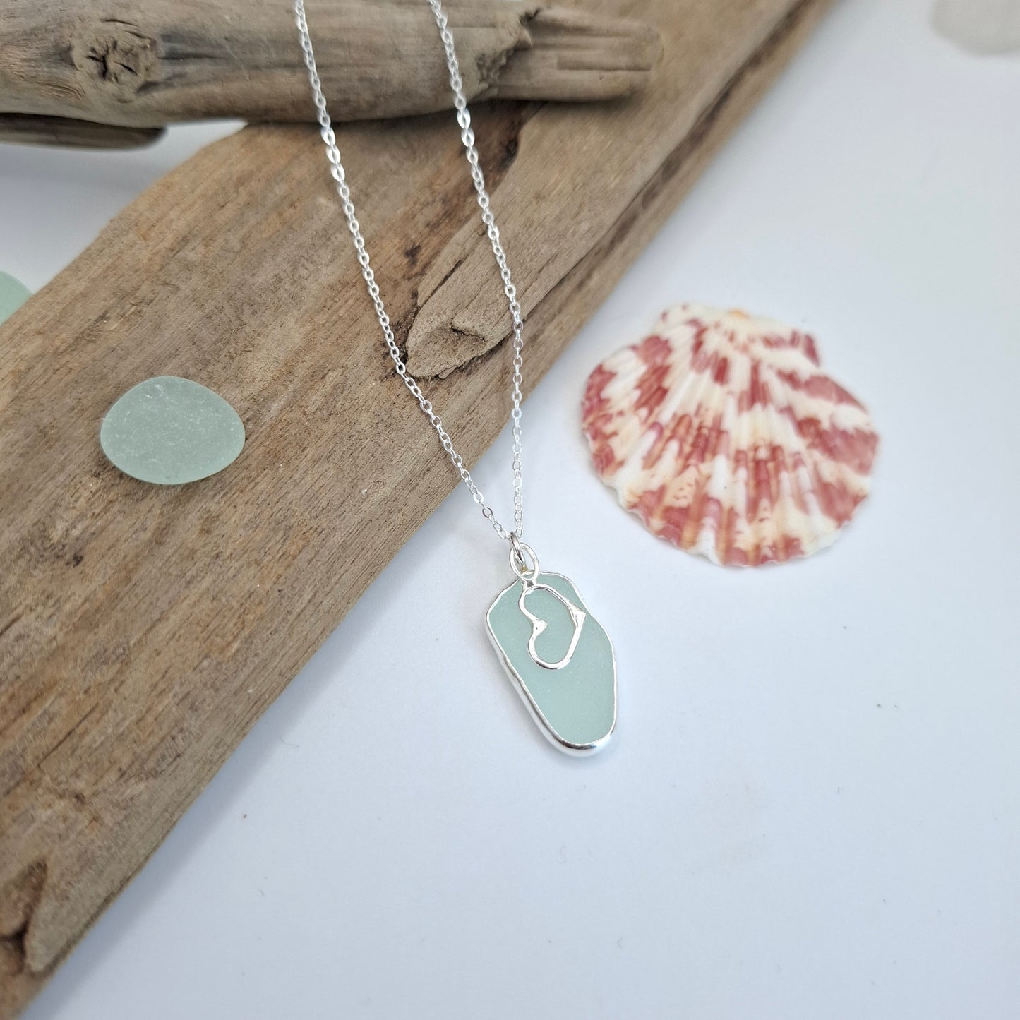 A silver necklace with a light green sea-foam stone pendant and a white heart outline, styled on a piece of driftwood with a seashell prop against a white background.