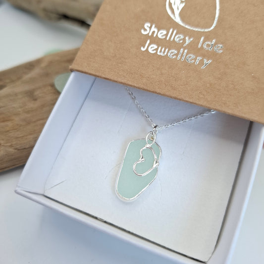 A silver necklace with an irregular light green sea glass pendant displayed in a white gift box next to a brown kraft card printed with 'Shelley Ide Jewellery'.
