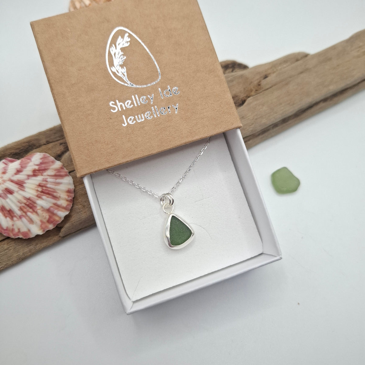 A silver necklace with a green teardrop-shaped stone pendant displayed in an open white jewelry box, with the kraft paper lid showing the Shelley Ide Jewellery brand. The box is styled on a white surface with a piece of driftwood, a seashell, and a small green stone.