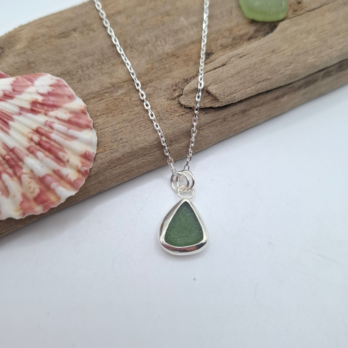 A silver chain necklace with a teardrop-shaped sea glass pendant in green, displayed on a piece of driftwood with a seashell prop.