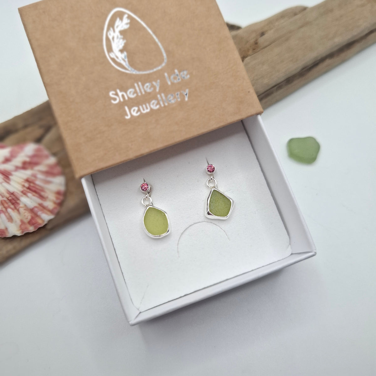 A pair of dangle earrings displayed in an open white jewelry box, with the kraft paper lid showing the brand name 'Shelley Isle Jewellery'. The earrings have irregular light green stone pendants in silver settings with small pink beads at the top.