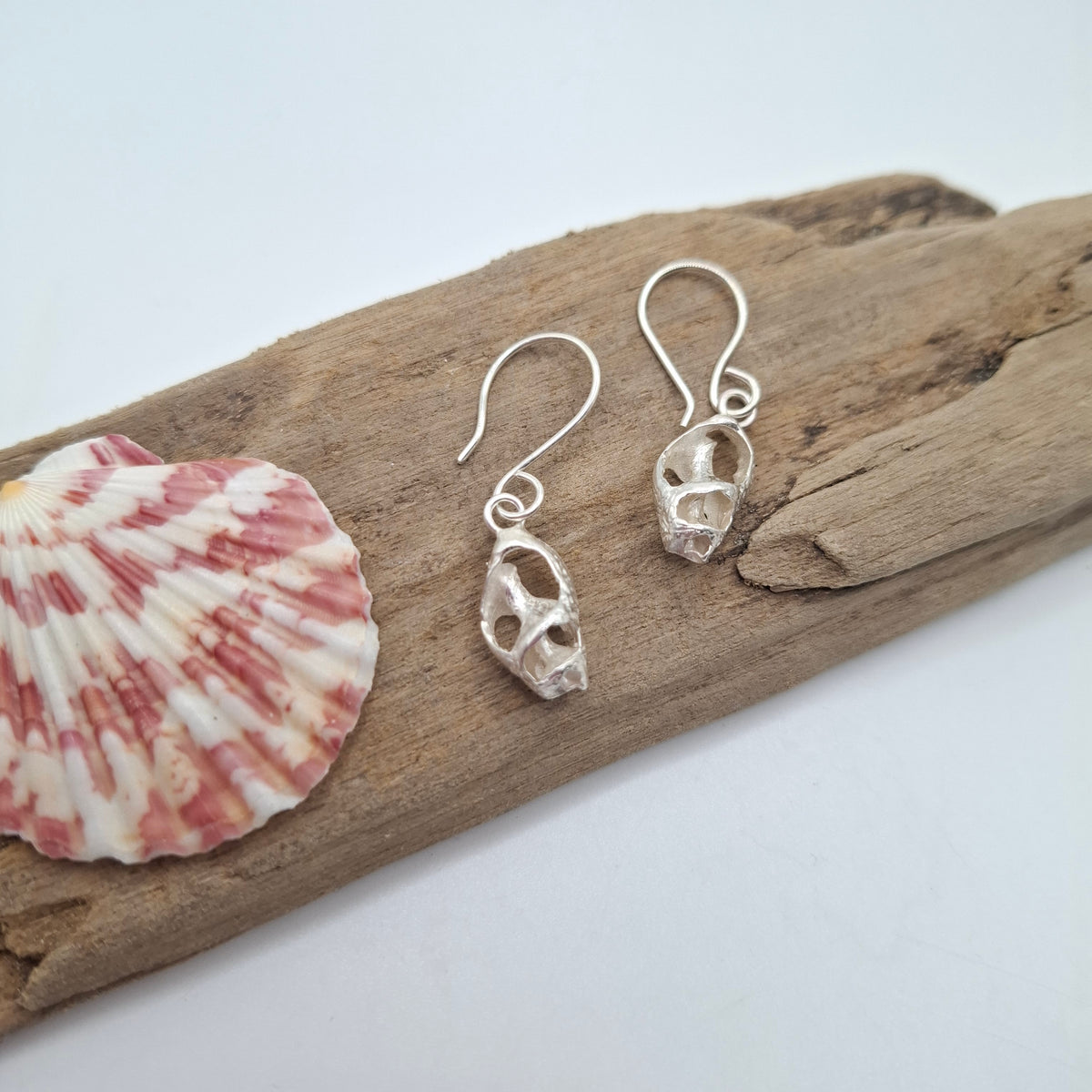 A pair of silver skull drop earrings displayed on a piece of driftwood next to a pink and white seashell against a clean white background.