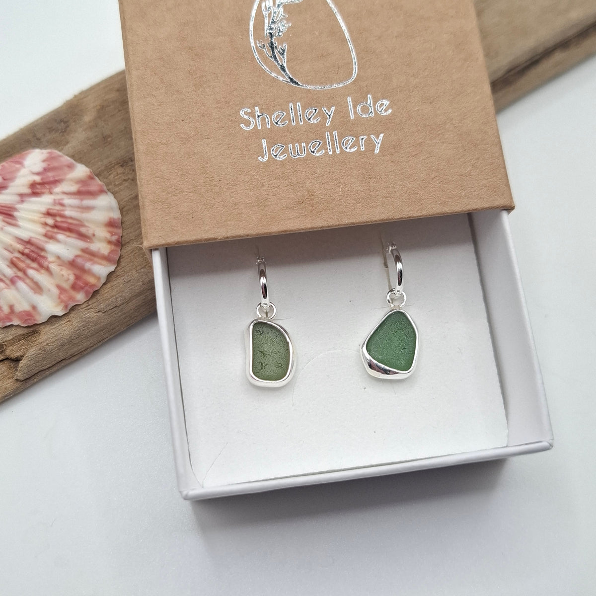 A pair of silver drop earrings with irregular green stone pendants displayed in an open white jewelry box, with a brown kraft lid printed with 'Shelley Ide Jewellery.' A seashell and driftwood are placed beside the box on a light surface.