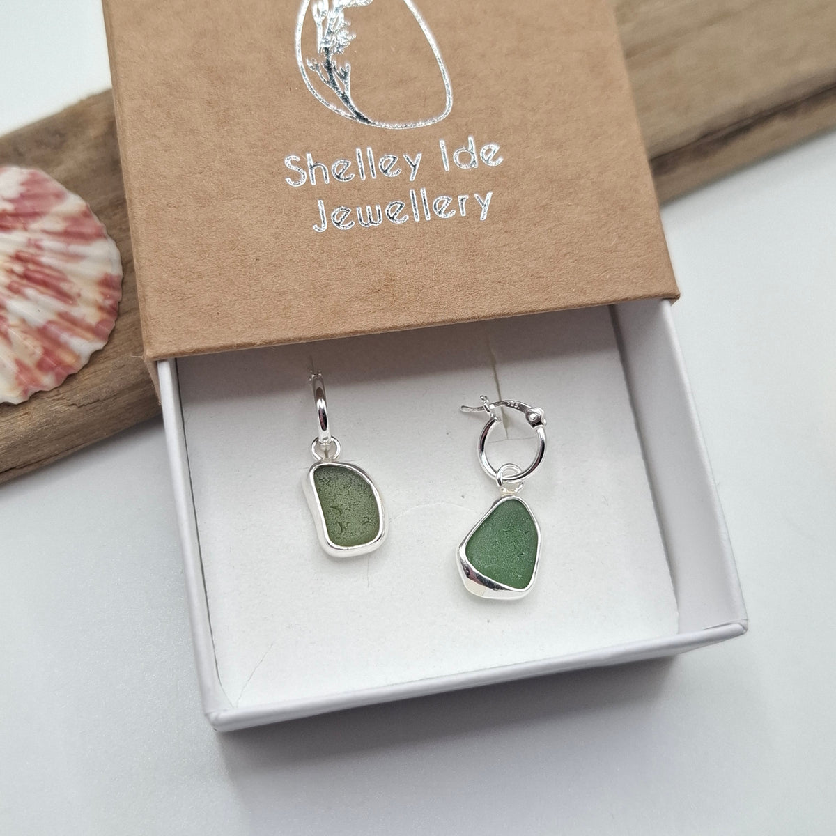 A pair of silver hoop earrings with irregular green stone pendants displayed in an open white jewelry box. The kraft paper lid is branded 'Shelley Ide Jewellery' and a seashell sits on a wooden board in the background.