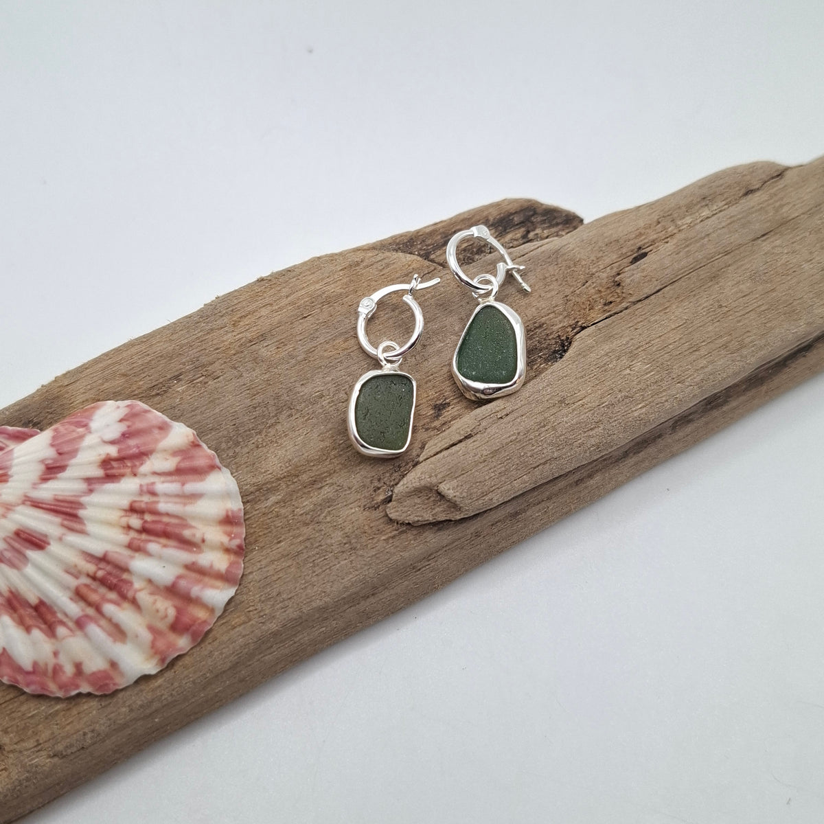 A pair of silver hoop earrings with rough green sea glass pendants, displayed on a piece of driftwood next to a pink and white seashell against a white background.