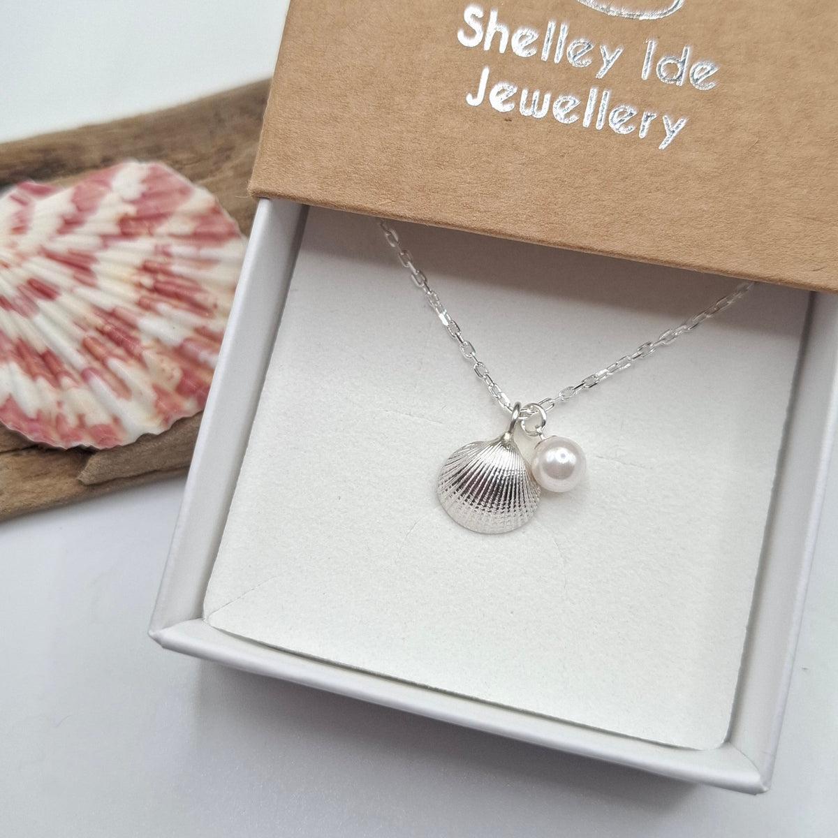A silver shell and pearl pendant necklace displayed in an open gift box with a kraft lid printed with the brand name 'Shelley Ide Jewellery'.