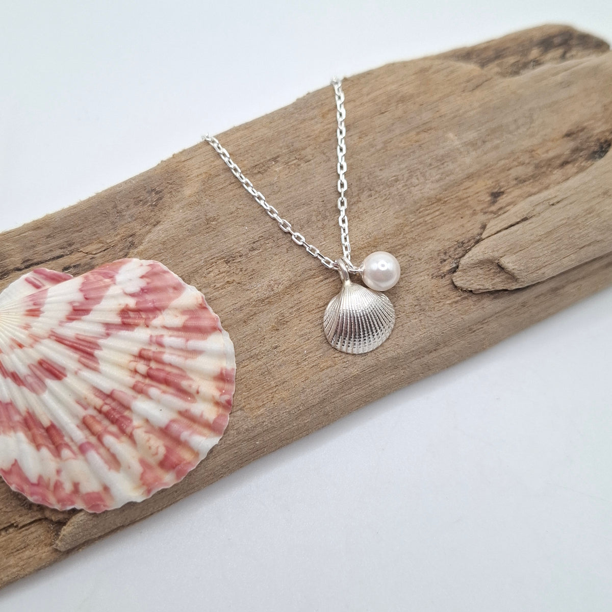 A silver necklace with a small ribbed seashell pendant and a white pearl bead, displayed on a piece of driftwood next to a pink and white seashell against a white background.