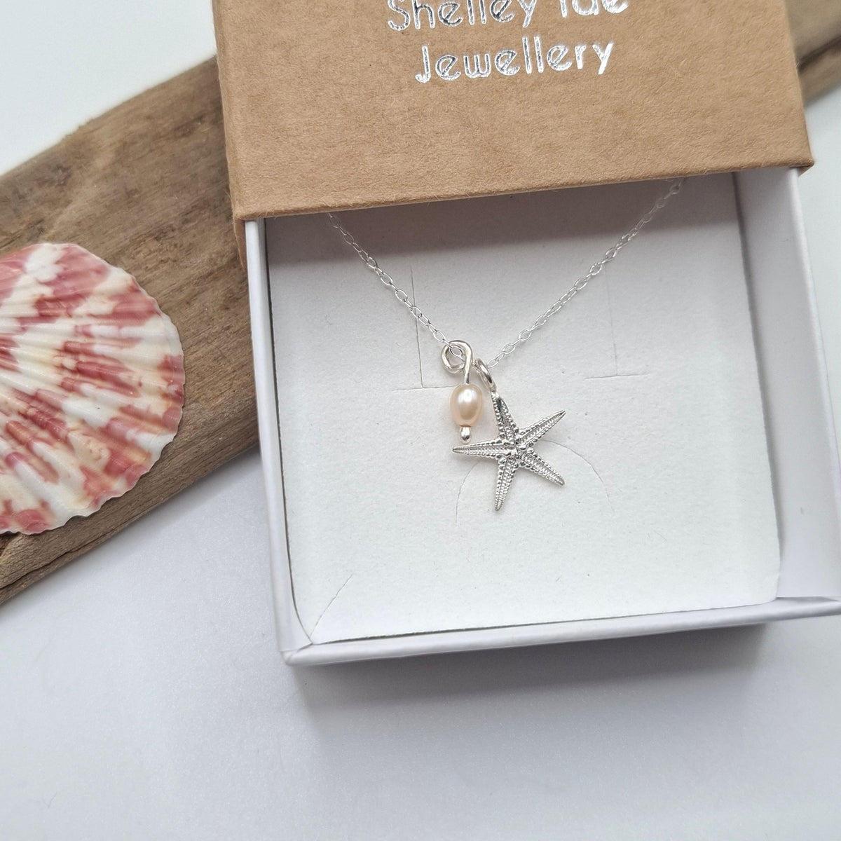 A silver starfish and pearl necklace displayed in an open white jewelry box with a brown kraft lid, styled with a seashell and driftwood on a light surface.