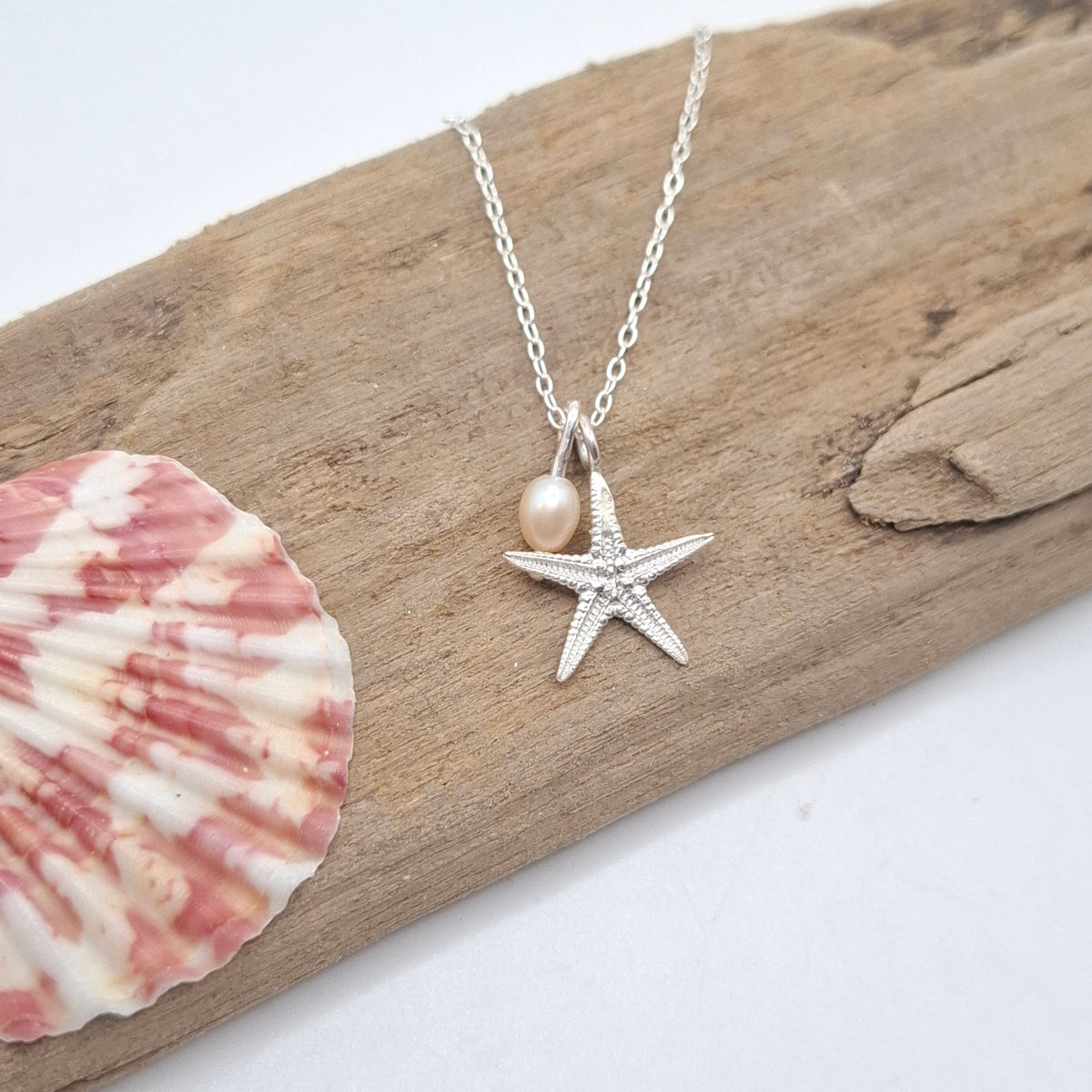A silver starfish pendant necklace with a small pearl bead, displayed on a piece of driftwood next to a pink and white seashell.