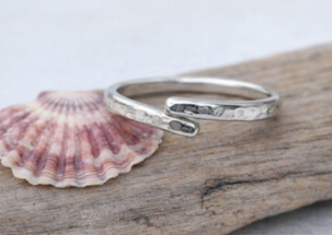A silver open band ring with a hammered texture, resting on a piece of driftwood next to a pink and white seashell.