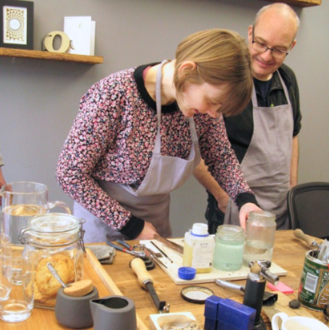 A woman in a floral long-sleeve top and white apron is leaning over a wooden worktable, mixing or pouring something into a glass jar. A man in a dark shirt and gray apron stands behind her.