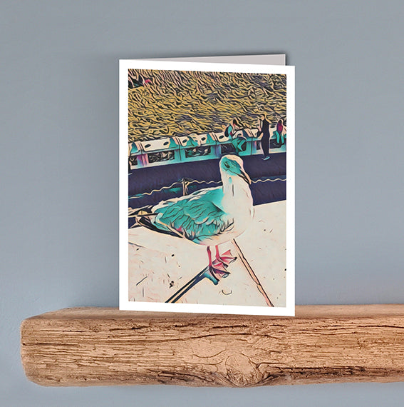 A colourful artistic print on a blank greeting card of a seagull standing on a wall with people in the background.